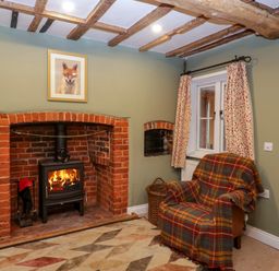 A room with a brick fireplace with a wood stove fire armchair with a plaid blanket window with curtains and wooden ceiling beams at 2 Loxter Villas in Wellington Heath near Ledbury