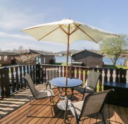 A wooden deck with a round table four chairs and a large umbrella overlooking lake cabins at Sunset Retreat South Lakeland Leisure Village near Carnforth