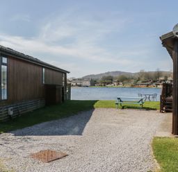 A gravel driveway between wooden buildings with a picnic table and chairs near a lake at Sunset Retreat in South Lakeland Leisure Village near Carnforth