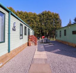 An outdoor area between two light green lodges with a paved walkway gravel and patio furniture at Lodge 2 in Silverdale