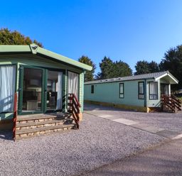 Two green mobile homes with steps in front on a gravel area with trees in the background at Lodge 3 in Silverdale