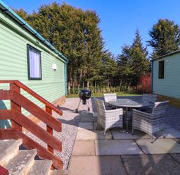 An outdoor patio area with a round table and four chairs between two light green lodges surrounded by trees at Lodge 3 in Silverdale