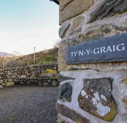 Stone wall with a sign reading TY'N-Y-GRAIG and a stone fence with plants and hills in the background at Tyn Y Graig in Fairbourne