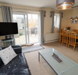 A living room with a black leather sofa and a glass coffee table next to a dining table with wooden chairs and a TV mounted near sliding doors at Sea Shore in Seahouses