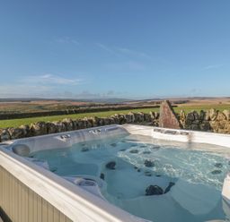 An outdoor hot tub next to a stone wall with fields and hills in the background at Petty Knowes Cottage in Rochester near Otterburn