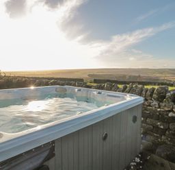 An outdoor hot tub near a stone wall with rolling hills in the background at Petty Knowes Cottage Rochester near Otterburn