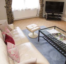 A living room with a cream sofa and patterned cushions a glass coffee table with magazines a cream ottoman a television on a stand floral curtains and beige carpet at Bank View in Chirnside