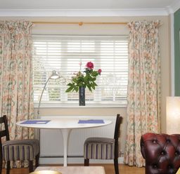 A dining area with a white oval table and two striped chairs by a window with floral curtains and a vase of flowers on the windowsill at Alway in Lifton