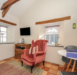 A sitting area with a pink armchair a wooden cabinet and a wall-mounted tv near windows and a table with chairs at The Barn in Lifton
