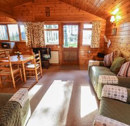 A wooden cabin living room with green upholstered sofas a round wooden dining table and kitchen appliances at Goldcrest in Withern near Alford