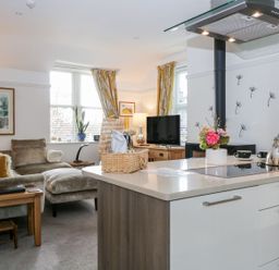 An open plan kitchen and living room with a kitchen island a stovetop a kettle and a flower vase in the kitchen and a sofa a TV and windows with curtains in the living area at The Bolt Hole in Kendal