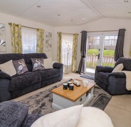 A living room with dark sofas, a wooden coffee table with mugs and a teapot, floral curtains, and glass doors at 98 The Heron in New Quay