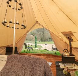 Inside a bell tent with a bed covered by a blanket a wood stove with a kettle and two chairs outside on grass at Bell tent 2 in Camerton near Timsbury