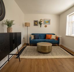 A living room with a blue sofa cushions round coffee table black cabinet wall art and a window at The Cove in Kessingland