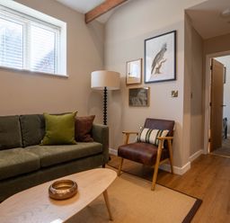 A living room with a green sofa pillows and a floor lamp next to a brown armchair with framed pictures on the wall at Dunes Cottage in Kessingland