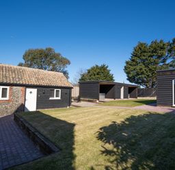 A backyard with a lawn, a brick path, a small building with tiled roof, and black wooden structures at Dunes Cottage in Kessingland