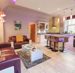 An open living room and kitchen with two sofas a glass coffee table a kitchen island with four stools and kitchen appliances at 1 The Old Coach House in Ambleside