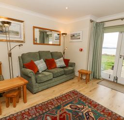 A sitting room with green sofa red and check cushions wooden tables floor lamps and glass doors opening to a garden at Carningli Suite near Newport