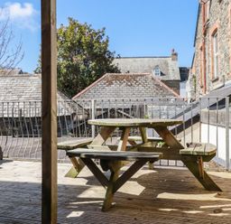 A wooden picnic table on a deck with metal railing and stone buildings in the background at St Georges in Padstow