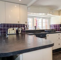 A kitchen with dark countertops white cabinets purple tiled backsplash and appliances at St Saviours in Padstow