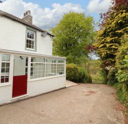 Exterior view of a white house with a red door and glass porch next to a driveway and trees at Kirkland Howe Cottage in Arlecdon