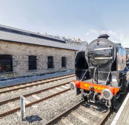 A steam locomotive at a train station platform with a stone building alongside Duchess of Hamilton @ Engine Shed in Whitby