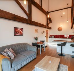 A living area with a gray sofa, a wooden table, two chairs, a kitchen island with two bar stools, and exposed wooden beams on the ceiling at Duchess of Hamilton @ Engine Shed in Whitby