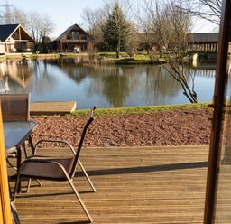 A wooden deck with chairs and table overlooking a pond with wooden houses around at The Green Monkey in North Hykeham