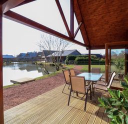 A covered wooden deck with a table and six chairs overlooking a pond and wooden houses at The Green Monkey in North Hykeham