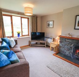 A living room with a brown sofa with blue cushions a stone fireplace a television on a stand and a window with curtains at Low Wood View in Hartsop