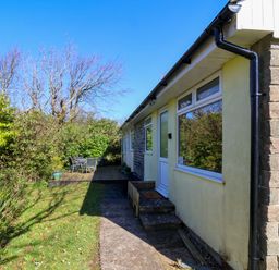 Exterior view of a single-story house with yellow walls and large windows next to a garden with trees and outdoor furniture at 24 The Glade in Kilkhampton