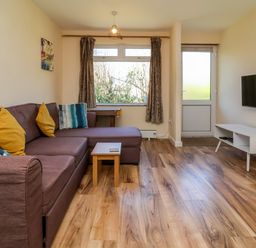 A living room with a brown sectional sofa and yellow pillows a small wooden table a wall mounted television and a window at 24 The Glade in Kilkhampton