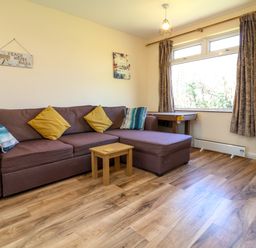 A living room with a brown sectional sofa with cushions a small wooden table wooden flooring and a window with curtains at 24 The Glade in Kilkhampton