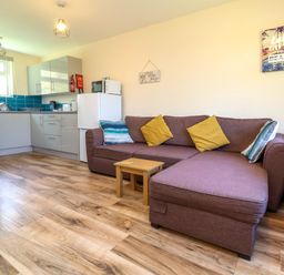 A living room with a brown sofa and cushions next to a small wooden table and an open kitchen area with grey cabinets and a white refrigerator at 24 The Glade in Kilkhampton