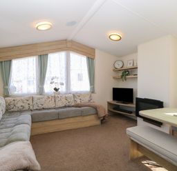 A living room with an L-shaped sofa, a TV on a wooden stand, shelves with decorations, and a dining table with a bench at 31B Foxburrow Hang in Fritton near Belton Norfolk