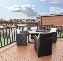 An outdoor patio with black wicker chairs with white cushions and a glass table surrounded by wooden buildings at Blossom Vale Lodge in Evesham