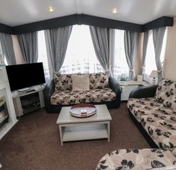 A living room with two floral sofas a TV a fireplace a coffee table and large windows with curtains at Rose Bank D8 in Primrose Valley