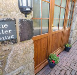 A stone wall with a sign reading The Baler House next to wooden doors with glass panels and pots with flowers on the ground at The Baler House in Hayle