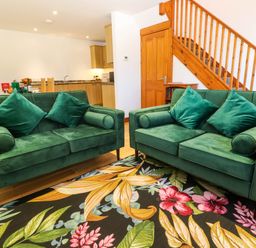 A living area with two green sofas on a floral rug near a wooden staircase and kitchen at The Baler House in Hayle