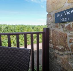 A brown wicker chair and wooden fence next to a stone wall with a Bay View Barn sign with greenery in the background at Bay View Barn in Hayle