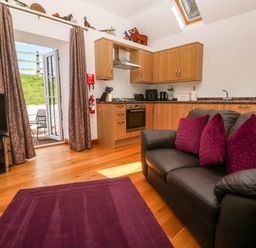 A living area with a black sofa and purple cushions near a kitchen with wooden cabinets and a TV at The Coach House in Hayle
