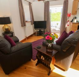 A living room with two black leather sofas a wooden table with flowers a lamp a television and kitchen area at The Coach House in Hayle