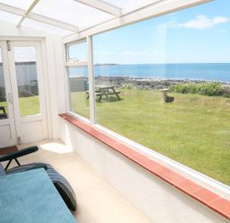 A sunroom with a cushioned chair and table overlooking a lawn with a picnic bench and ocean view at Tegawelon in Llandanwg near Harlech