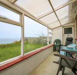 A sunroom with plastic and reclining chairs and large windows showing a grass lawn and a stone wall at Tegawelon in Llandanwg near Harlech