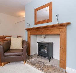 A living room with a brown armchair a wooden dining table and chairs a clock on the wall a fireplace with a wooden mantel and a black stove inside at Spring Cottage in Birdlip near Brockworth