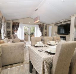 A dining table with tableware and beige chairs next to a living area with sofas and large glass doors at Water's Reach White Cross Bay Holiday Park near Troutbeck Bridge