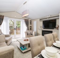 A living room with beige sofas a coffee table dining chairs with place settings a television and glass doors to a balcony at Water's Reach White Cross Bay Holiday Park near Troutbeck Bridge