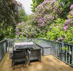 An outdoor deck with wicker furniture a table and a storage bench surrounded by blooming plants at Serenity Skiptory Howe 10 White Cross Bay near Windermere