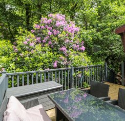 An outdoor patio with a glass table and wicker chairs surrounded by green plants and pink flowers at Serenity Skiptory Howe 10 White Cross Bay near Windermere
