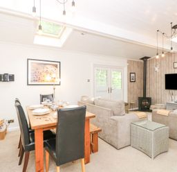 A living and dining area with a wooden dining table set with plates and glasses and beige sofas around a TV mounted on the wall at Serenity Skiptory Howe 10 White Cross Bay near Windermere